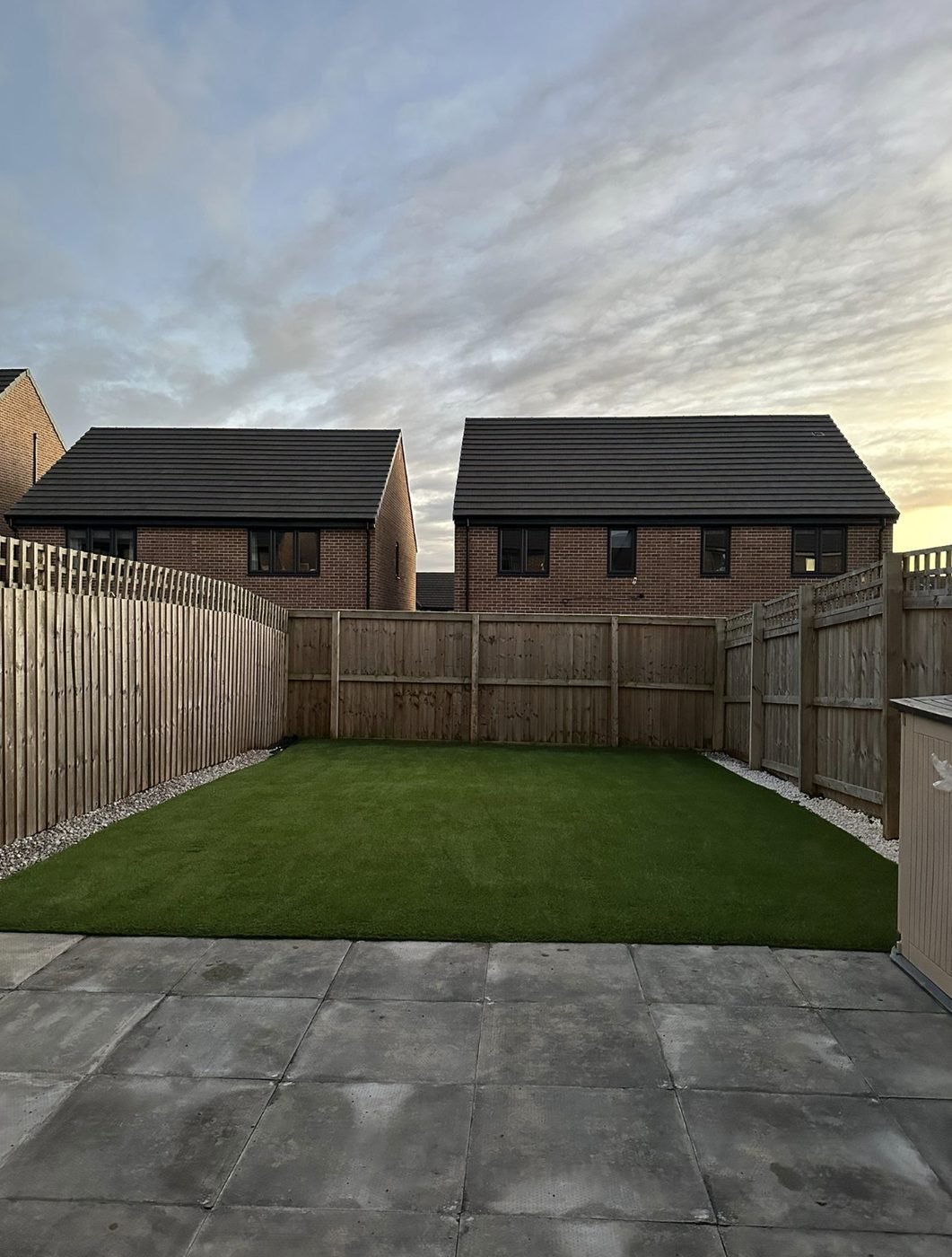 Artificial grass lawn with gravel margins in a new build garden at dusk, with a paved patio in the foreground