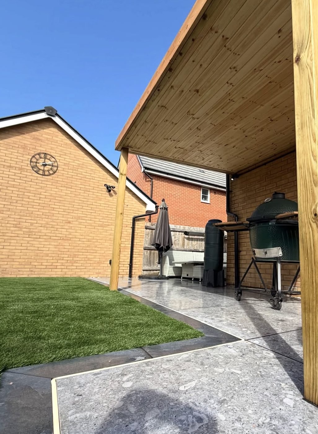 Timber canopy over a paved outdoor kitchen area with a ceramic barbecue, alongside an artificial grass lawn